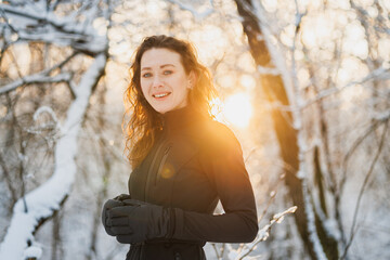 Positive woman in ski suit and gloves looking at camera in winter forest during sunset 