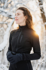 Curly woman in gloves and black ski suit standing in forest during sunset 