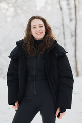 Brunette woman in ski suit and outerwear smiling at camera in winter forest 