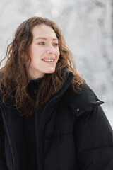 Portrait of curly woman in outerwear smiling in winter forest 