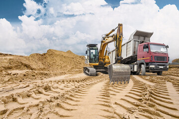 Obraz premium Deep tracks mark the sandy ground where heavy machinery works. A loader and truck move sand near a large pile, showing active construction in progress
