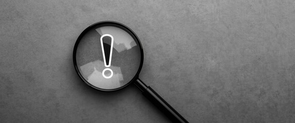 Magnifying glass in black and white focusing on exclamation mark against gray background, signaling alert and risk management