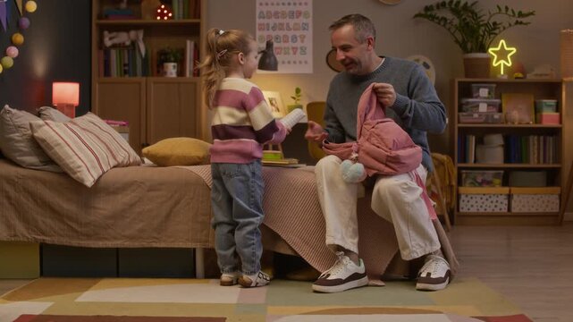 Smiling mature father sitting on bed edge and helping her little daughter to pack books and notebooks in her brand new backpack