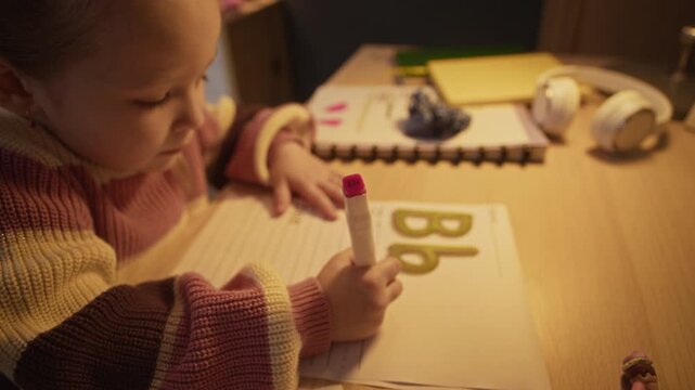 Pull out shot of little Caucasian kid sitting at wooden desk, using pink felt pen to practice her handwriting skills