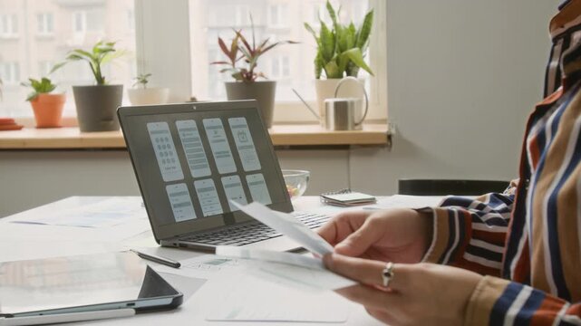 Medium side shot of unrecognizable female UI engineer in striped shirt examining cutouts of new wireframe prototypes, checking layout concepts on laptop screen, at desk by window in office