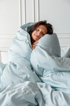 A woman is sitting in bed wrapped in light blankets. She is smiling as she enjoys her morning in a bright room. Plants are visible in the background with soft lighting.