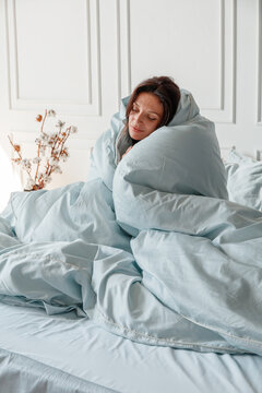 A woman is sitting in bed wrapped in light blankets. She is smiling as she enjoys her morning in a bright room. Plants are visible in the background with soft lighting.