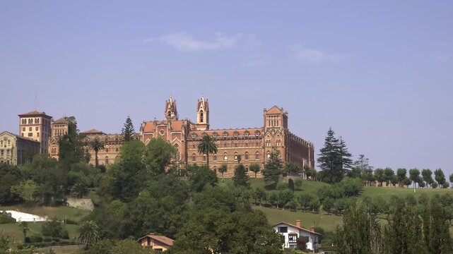 Scenic view of the historic Comillas Pontifical University building, an architectural marvel of Catalan modernism nestled on a lush green hill in Cantabria, Spain, during a sunny day