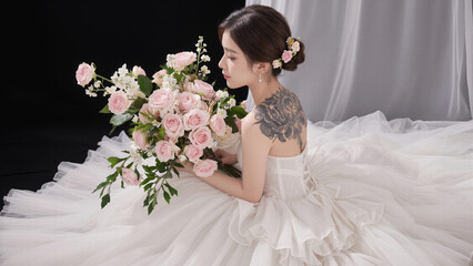 Romantic bridal portrait of a woman in a white wedding dress with a floral hair accessory and back tattoo holding a beautiful bouquet of fresh pink roses.