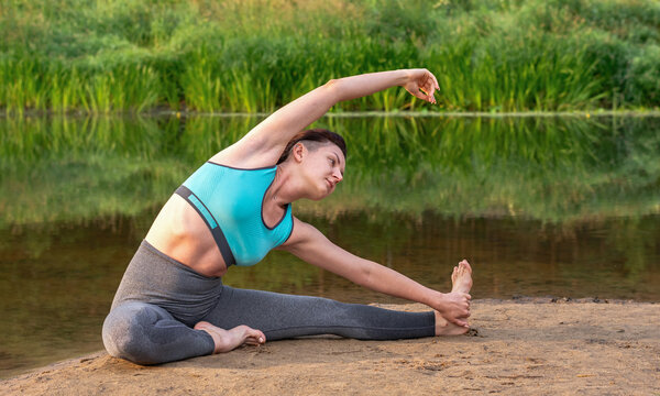 Young sporty woman practicing yoga by the river, doing Head-to-Knee Forward Bend (Janu Sirsasana).