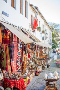 Vibrant outdoor souvenir market along a historic European street showcasing handmade rugs, patterned fabrics, vintage lanterns, metal crafts, and local artisan goods beneath awnings