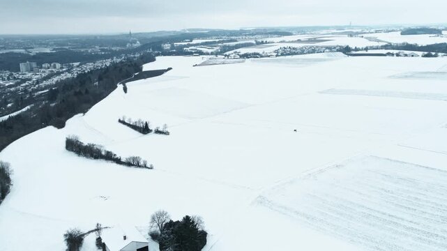 Forward drone flight above snow-covered fields near Elchingen, distant village, and winter horizon under overcast sky.