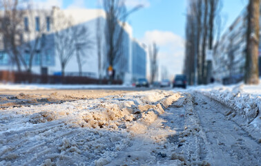 Obraz premium Snowy urban street with icy tire tracks and slush, cars driving past apartment buildings on a cold winter day. Low-angle view highlighting winter road conditions