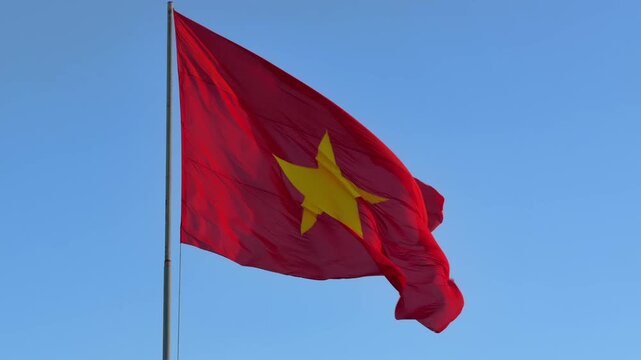 Slow motion Vietnam flag waving dynamically in the wind, illuminated by low evening sun with blue sky behind it during golden hour. Cultural heritage.
