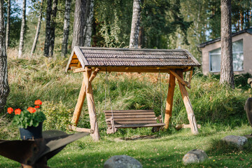 Wooden swing set with a thatched roof in a lush green garden surrounded by trees and a flower pot with red blooms in the foreground © Raivo
