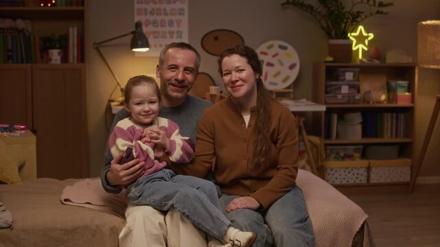 Portrait of smiling mother and father looking into camera and little daughter sitting on dads laps posing for camera