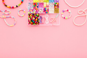 Bracelet, necklaces, scissors, elastic strings and case with colorful plastic parts on pink table background. Pastel color. Closeup. Creating beaded jewellery for child girl. Flat lay. Top down view.