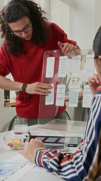Medium vertical shot of female project manager sitting at team meeting, listening to enthusiastic young gen Z male UI designer in red sweater, presenting wireframe concepts for new smartphone app