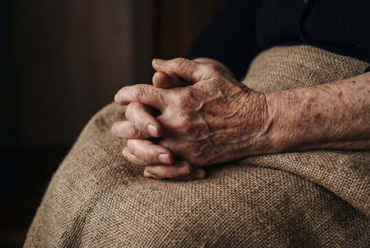 Wrinkled hands of senior woman clasped together resting on rough brown burlap sackcloth fabric