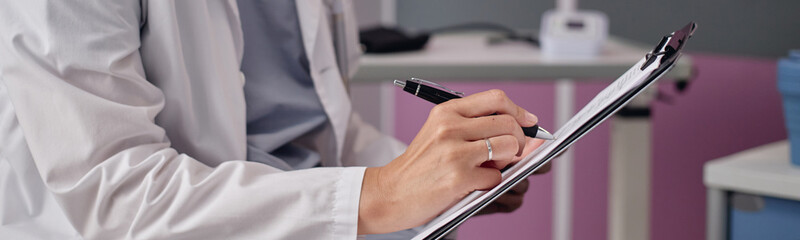 Website header shot of adult woman writing on clipboard with pen in medical office, wearing white coat, sitting at desk in vascular center, focusing on patient documentation and healthcare records © Seventyfour