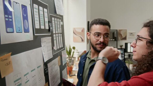 Side medium close-up of two cheerful young multiethnic male UX developers discussing wireframe concepts displayed on project board, brainstorming and suggesting usability improvements