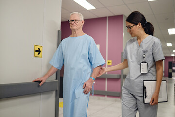 Senior Caucasian man walking in hospital corridor with assistance from young adult Asian woman...