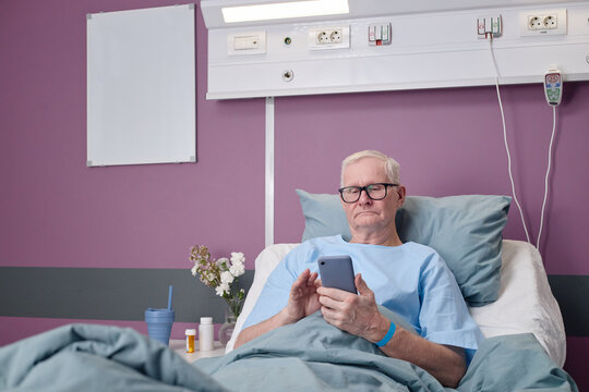 Senior Caucasian man lying in hospital bed using smartphone, wearing medical gown and glasses, recovering in vascular center patient room with medication and flowers on bedside table