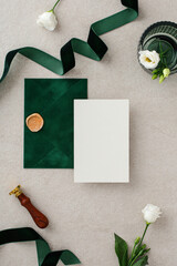 Wedding stationery set. Dark green envelope, ribbon, blank invitation card mockup, wax seal stamp, and white flowers on stone background.