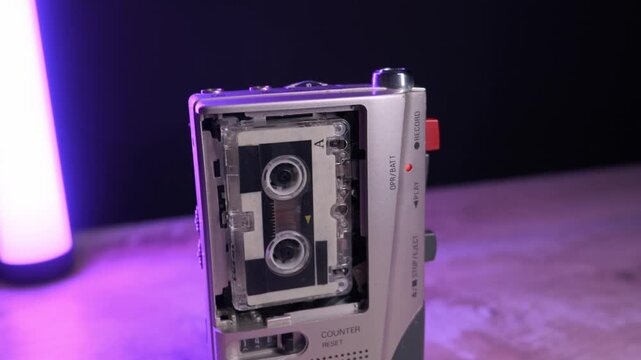 Silver microcassette recorder records audio with spinning reels against changing neon lights on a wooden table. Concept of espionage, retro technology, 80s nostalgia, and lo-fi audio recording.