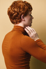 Redhead young man in rust turtleneck sweater posing sideways with hand near neck on beige background. Profile portrait of fashionable male with curly hair and casual style indoors. © SHOTPRIME STUDIO