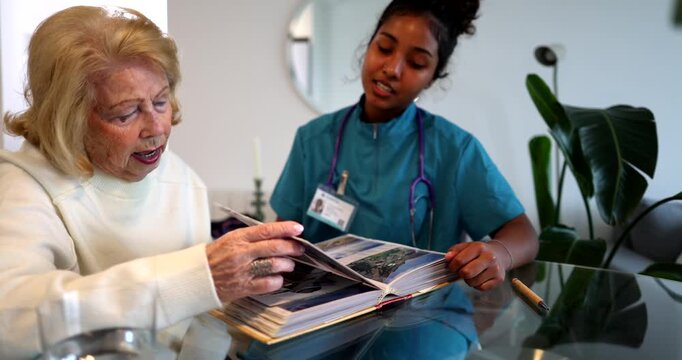 Senior woman sitting at a table with her caregiver, looking together at old pictures in a photo album, reminiscing about memories from the past during a home care assistance visit