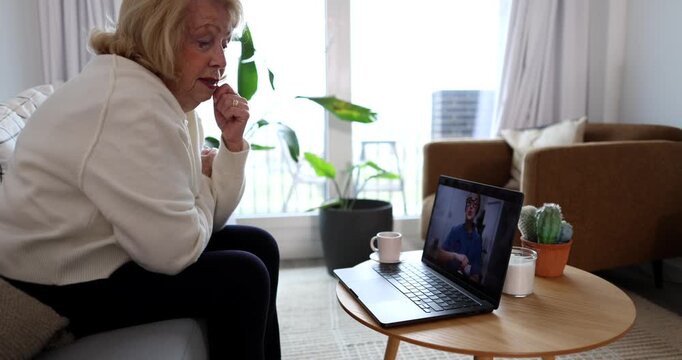 Senior woman sitting in her living room, actively engaged in an online video consultation with a male therapist on a laptop, discussing her mental health and receiving remote support