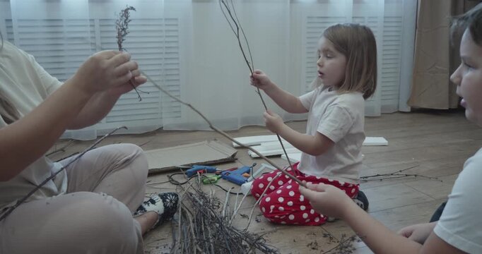 Family sitting on the floor crafting with natural tree branches and a hot glue gun, enjoying cozy weekend quality time while kids develop fine motor skills and creativity.