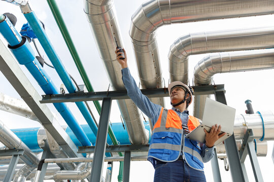 Engineer in safety uniform inspects industrial pipelines using laptop and walkie-talkie at facility, ensuring proper operation, system monitoring in an industrial.