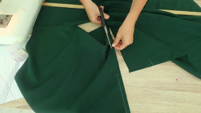 A person carefully cuts deep green fabric with scissors on a wooden table beside a sewing machine, precision tailoring in a well lit craft space.