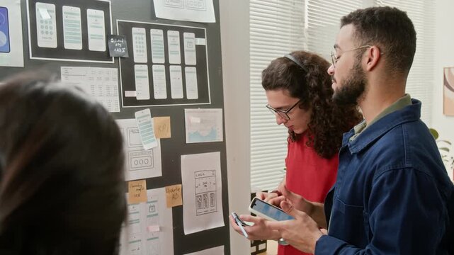 Medium close-up of diverse UX design team reviewing mobile application wireframes at brainstorming meeting by wall display board in office, debating layout, functionality and usability options