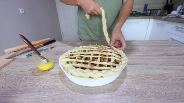 A baker weaves thin dough strips over a filled apple pie in a lattice pattern, meticulous, hands-on finishing for a classic homemade dessert.