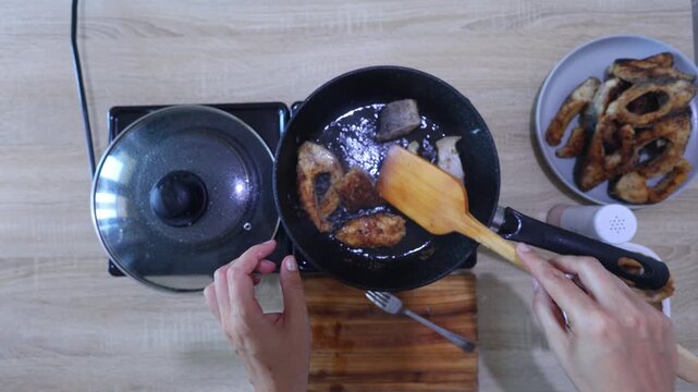 Overhead view: hands place floured fish steaks into a hot oiled pan for frying, batch cooking with finished pieces already plated nearby.