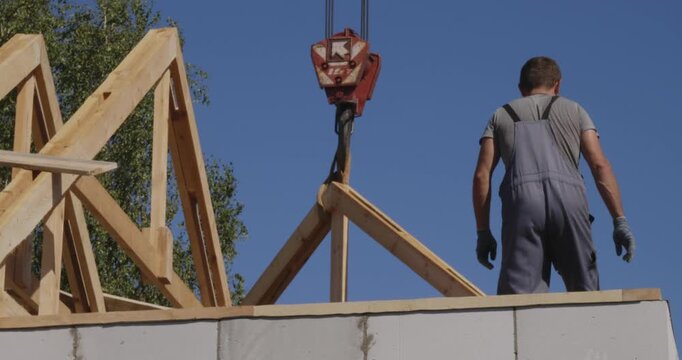 Professional builder on a construction site guiding a wooden roof truss being lowered by a crane, preparing to install the new timber frame structure on a cinder block house.
