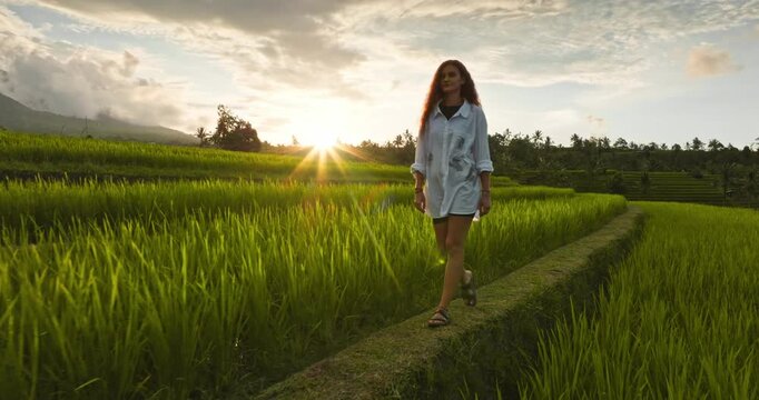 People enjoy Bali nature. Woman walking narrow path on vibrant green rice paddies at sunset with tropical landscape and distant mountains, embodying tranquility and sustainable travel in Indonesia
