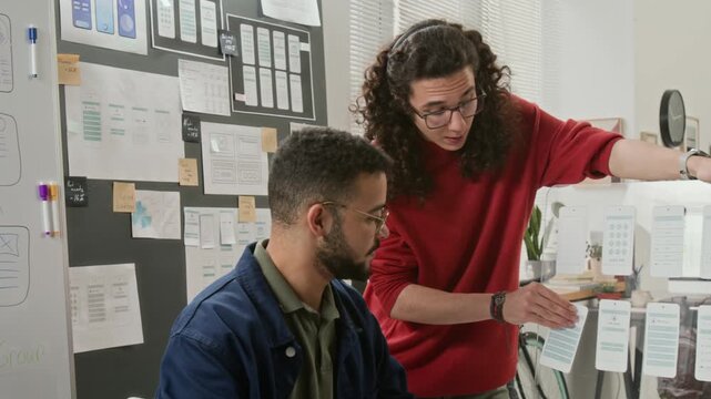Medium shot of diverse male and female UI designers discussing user experience scenarios while examining wireframes for new mobile app project at brainstorming meeting to improve usability