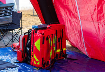 Red medical backpacks and emergency bags with reflective elements placed on a blue tarp in front of a red operational tent during rescue drills. © Adam