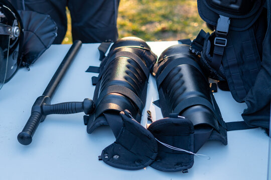 Protective gear for correctional officers: black shin guards and a tonfa baton displayed on a table during a presentation of riot control and transport equipment.