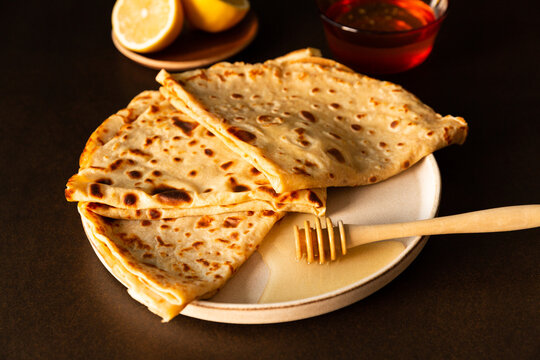 Gros plan sur des cr&ecirc;pes pli&eacute;es en triangle et pos&eacute;es en &eacute;ventail sur une assiette avec une cuill&egrave;re &agrave; miel en bois au premier plan, citron coup&eacute; et coupelle de miel en arri&egrave;re-plan, lumi&egrave;re dor&eacute;e