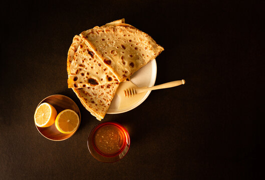 Trois cr&ecirc;pes pli&eacute;es en triangle et pos&eacute;es en &eacute;ventail sur une assiette avec une cuill&egrave;re &agrave; miel, citron coup&eacute; en deux et coupelle en verre remplie de miel, vue du dessus, fond brun fonc&eacute;, lumineux
