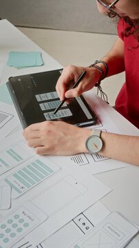 High-angle vertical shot of androgynous curly young user interface designer in red sweater choosing wireframes on touchscreen tablet while creating new mobile application in office