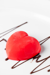 A glossy red heart-shaped mousse cake sits on a white plate with a chocolate drizzle. The focus is sharp on the dessert, suggesting love, romance, and Valentine's Day.