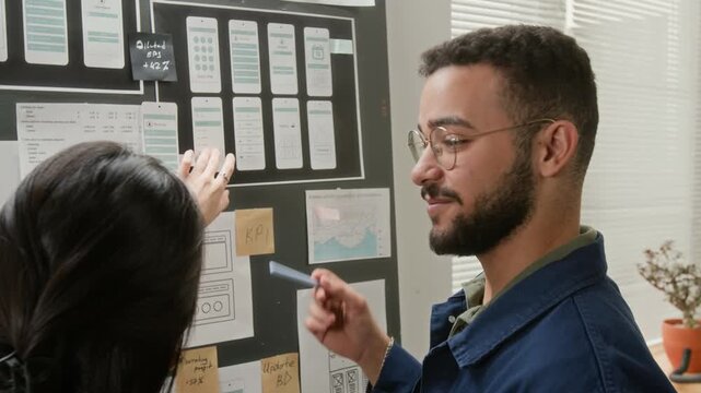 Rear shot of cheerful polyethnic male UX developer presenting various iterations of wireframe concepts to Hispanic female project manager, while working on mobile e-commerce application
