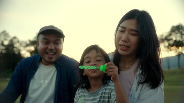 Parents and their young son blowing soap bubbles together sitting yard at the park during sunset, people joyful family bonding moment filled with laughter playful and childhood in happy family time.
