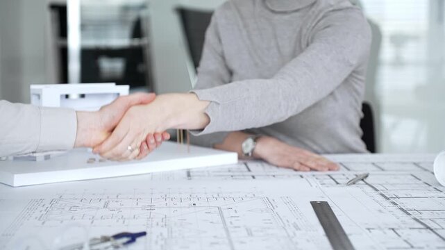 Handshaking between two architects finalizing a construction project deal, business partners agreeing on engineering blueprints and architectural models in an office, close up